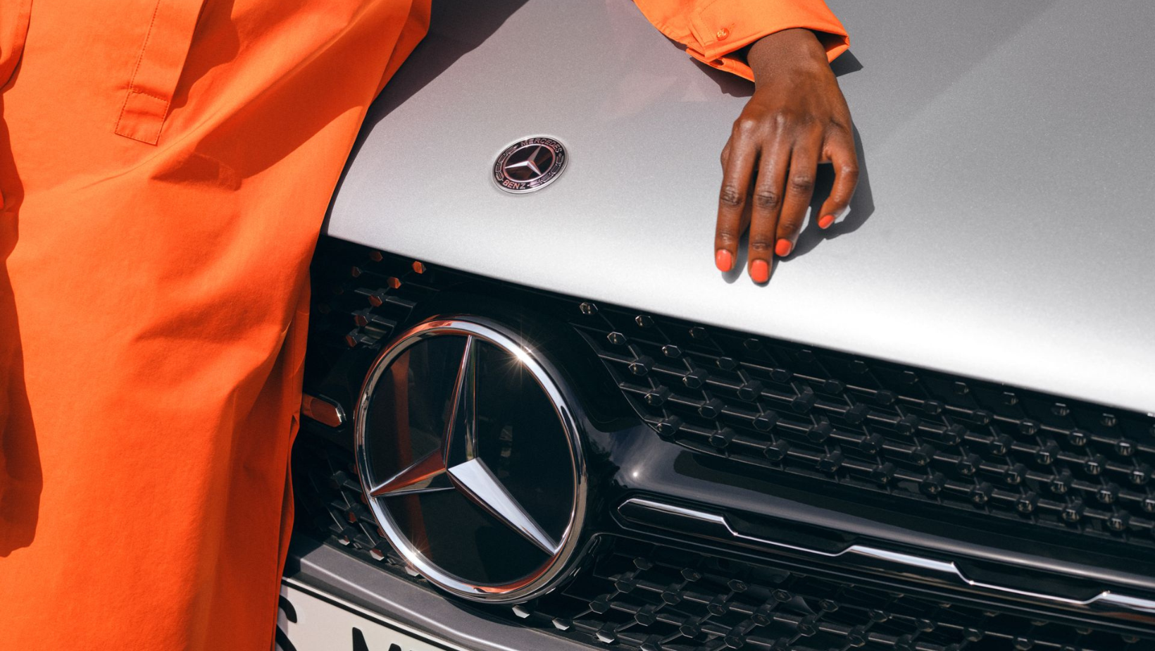 La photo met en avant l'avant d'un véhicule Mercedes-Benz, mettant en évidence le logo emblématique de la marque. Sur le capot, repose ce qui semble être le buste d'une femme, avec sa main délicatement posée à proximité du logo. Elle porte une tenue orange assortie à son vernis à ongles. Cette composition crée une esthétique captivante, mélangeant le design automobile de luxe avec une touche artistique et glamour, tout en mettant en valeur le logo emblématique de Mercedes-Benz.